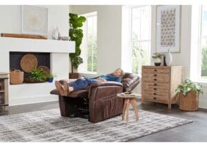 A person relaxes on a Dione Medium - Lift Chair by Golden Technologies in a modern living room with large windows, light wood furniture, a gray patterned rug, and green plants.