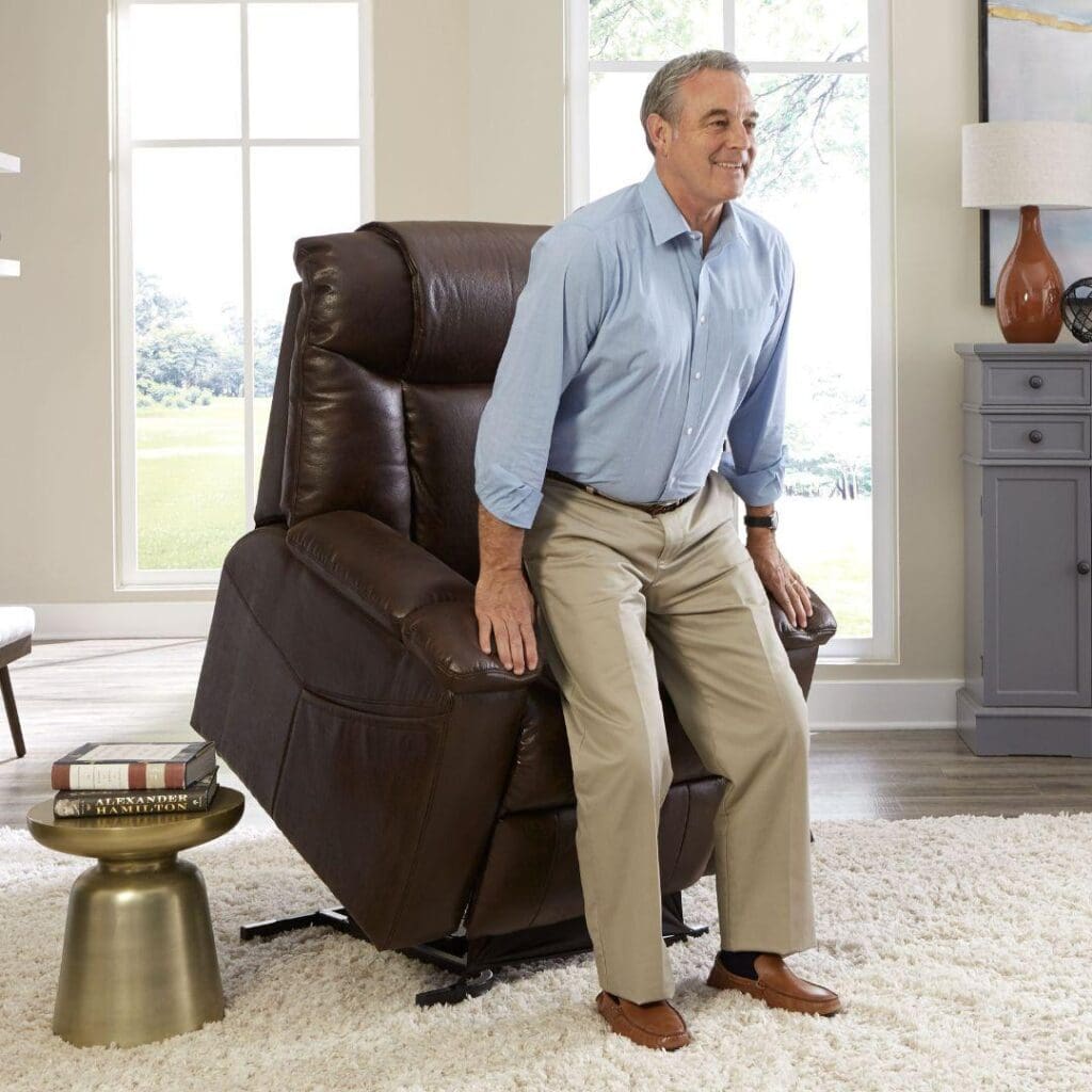 An older man in a blue shirt and khaki pants uses the Rhea Large - Lift Chair by Golden Technologies to stand up in a bright living room with large windows, a gray cabinet, and a small round table.