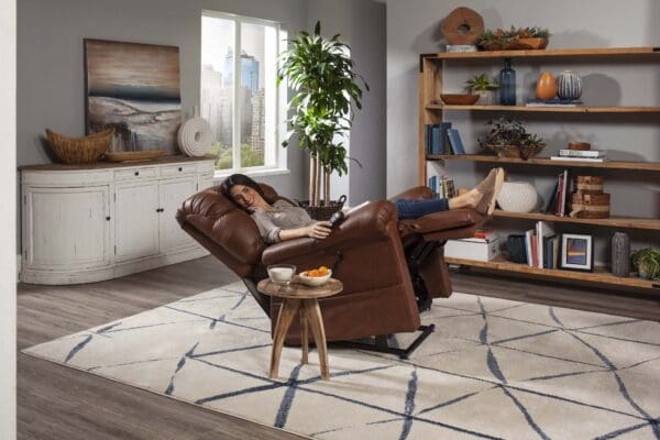A woman relaxes with her feet up in a Cloud Medium Large - Lift Chair by Golden Technologies, holding a remote in a cozy living room with bookshelves, a white cabinet, and a small round table with snacks.