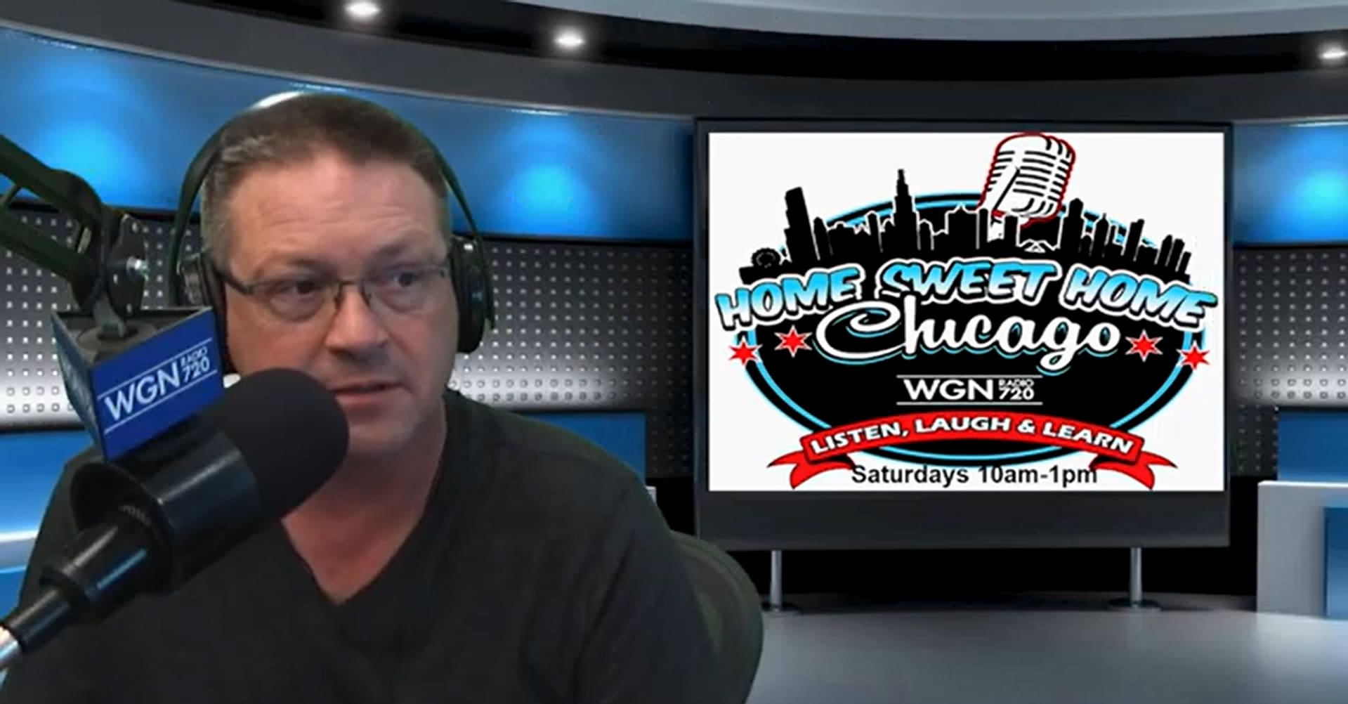 A man wearing glasses and headphones speaks into a microphone in a radio studio. Behind him is a large screen displaying the Home Sweet Home Chicago radio show logo and broadcast details.