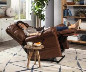 A woman relaxes and smiles with a remote in hand in her cozy living room, seated in the Cloud with Twilight Small/Medium Lift Chair by Golden Technologies. A small table with fruit and a drink is beside her, framed by shelves and a window.