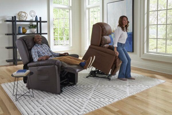 A man relaxes in a brown recliner while a woman smiles beside the Relaxer Large - Lift Chair by Golden Technologies as it rises upright in a bright, modern living room with large windows.