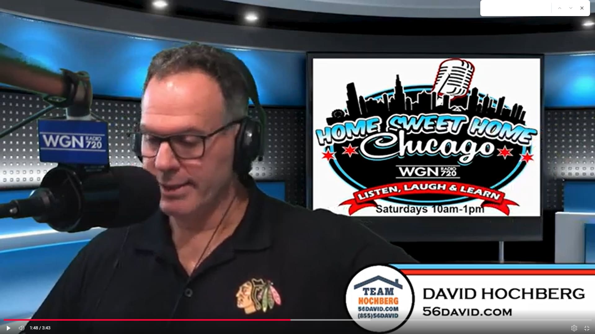 A man wearing glasses and headphones speaks into a microphone in a radio studio. Behind him, a screen displays the Home Sweet Home Chicago WGN 720 radio show logo and show details. A name tag reads David Hochberg.