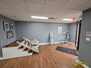 A small room with gray walls, two stair lift chairs on display, an exercise machine on a mat, informational pamphlets on a rack, framed information on the wall, and a wood-look floor. The Access company logo is visible.