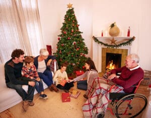 A family of six, including two children and an older man in a wheelchair, sits around a decorated Christmas tree exchanging gifts in a cozy living room with a lit fireplace.