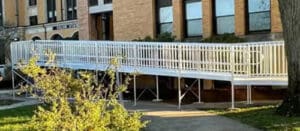 A long, white wheelchair-accessible ramp with railings leads to the entrance of a brick building, surrounded by grass and trees.