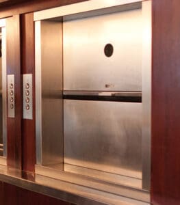 A Dumbwaiters & Material Lifts by Matot unit with closed stainless steel doors is set into a wooden wall, featuring control panels with buttons on both sides, highlighting efficient residential dumbwaiter design.
