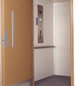 The Prolift Vertical Platform Lift by Savaria features an open ADA-compliant design with a beige door, white walls, metal wall rail, right-side control panel with buttons and speaker, gray floor, and overhead lighting for accessibility.