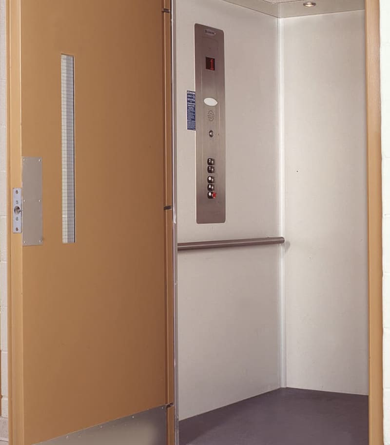The Prolift Vertical Platform Lift by Savaria features an open ADA-compliant design with a beige door, white walls, metal wall rail, right-side control panel with buttons and speaker, gray floor, and overhead lighting for accessibility.