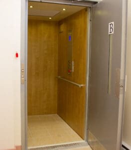 The Prolift Vertical Platform Lift by Savaria features a light wood interior, beige tiled floor, metal handrail, and control panel. The silver door displays a B sign on the adjacent wall. The lift is currently empty.
