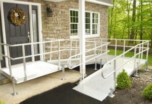 A house entrance with two aluminum wheelchair ramps and handrails, leading up to a front door with a wreath. The house has stone siding, large windows, and is surrounded by greenery.