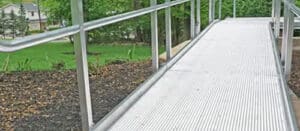 A metal wheelchair ramp with handrails on both sides, surrounded by mulch and green grass, leading to an outdoor area with trees and buildings in the background.