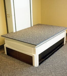 A white, rectangular pet crate with a carpeted top and dark tinted side panels sits on a carpeted floor by a beige wall, echoing the clean design of the Telecab Home Lift by Savaria.