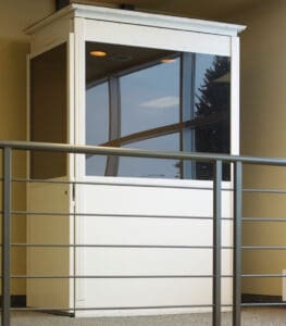 A small white security booth with large glass windows stands indoors near a metal railing, beside a Savaria Telecab Home Lift that reflects ceiling lights and offers a view of a tree outside.
