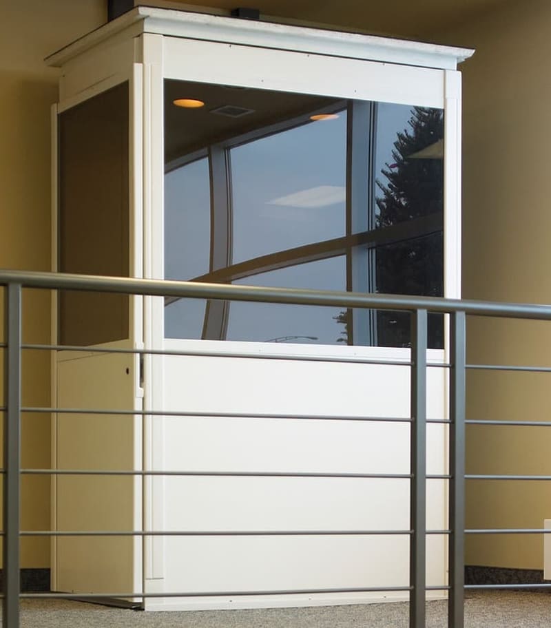 A small white security booth with large glass windows stands indoors near a metal railing, beside a Savaria Telecab Home Lift that reflects ceiling lights and offers a view of a tree outside. A small white security booth with large glass windows stands indoors near a metal railing, beside a Savaria Telecab Home Lift that reflects ceiling lights and offers a view of a tree outside.