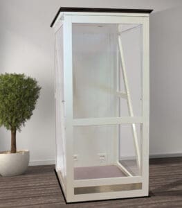 A white, glass-paneled phone booth stands indoors on a wooden floor, resembling the modern design of the Telecab17 Home Lift by Savaria, with a small potted tree nearby against a plain light-colored wall.