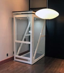 A compact Telecab17 Home Lift by Savaria, featuring glass walls and a white frame, is installed in a corner with wooden floors and minimal decor. A large round white hanging lamp nearby highlights its modern style.