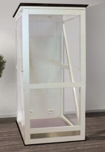 A white Telecab17 Home Lift by Savaria with glass walls stands indoors on a dark wood floor, resembling a compact elevator. A small plant sits to the left against a white wall, and electrical outlets are visible at the lift’s base.