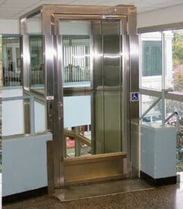 A compact V-1504 Lux Vertical Platform Lift by Savaria, featuring glass walls and a stainless steel frame, is installed indoors between two floors beside a staircase with blue railings and a visible disability symbol on the front.
