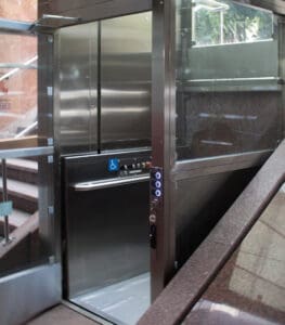 A modern glass and metal elevator with open doors, accessible buttons, and a wheelchair symbol—possibly a V-1504 Lux Vertical Platform Lift by Savaria—is positioned beside a staircase in a well-lit building.