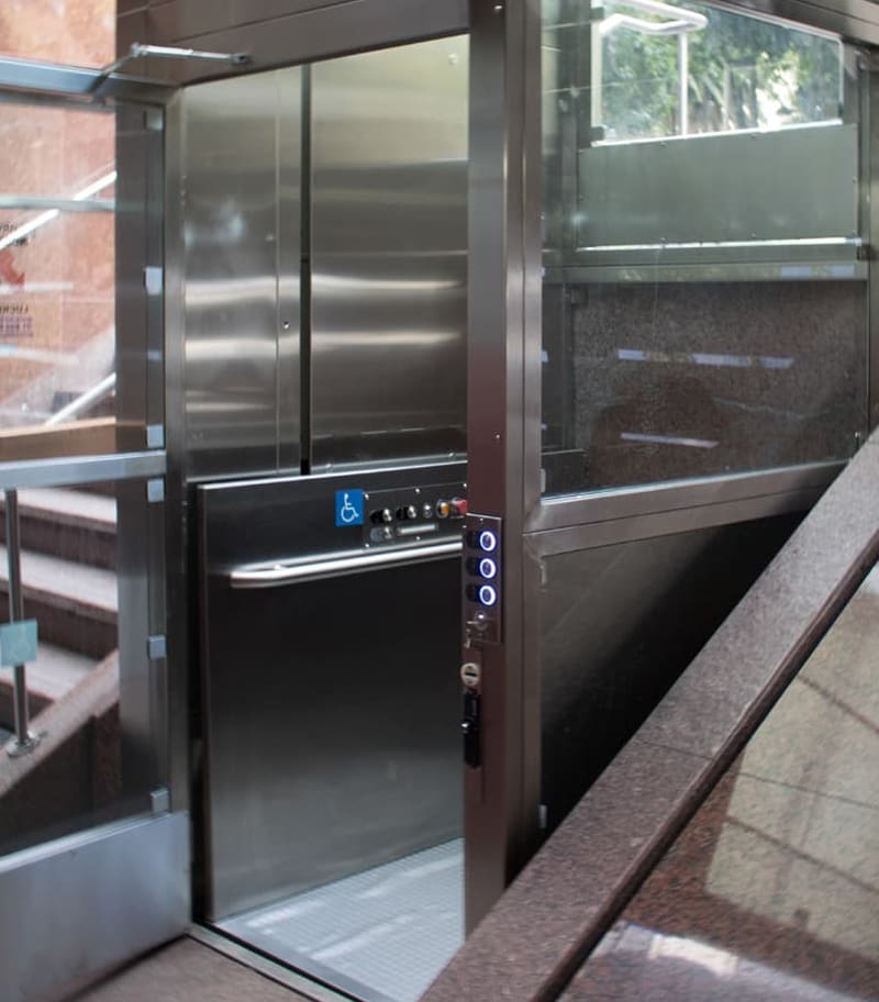 A modern glass and metal elevator with open doors, accessible buttons, and a wheelchair symbol—possibly a V-1504 Lux Vertical Platform Lift by Savaria—is positioned beside a staircase in a well-lit building.