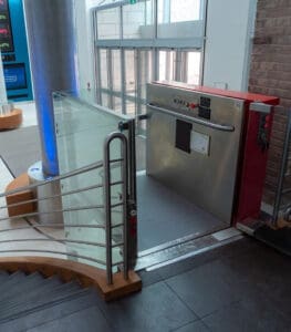 A Savaria V-1504 Prestige Vertical Platform Lift stands beside the stairs in a modern lobby, surrounded by glass walls, metal railings, and sliding glass entrance doors.