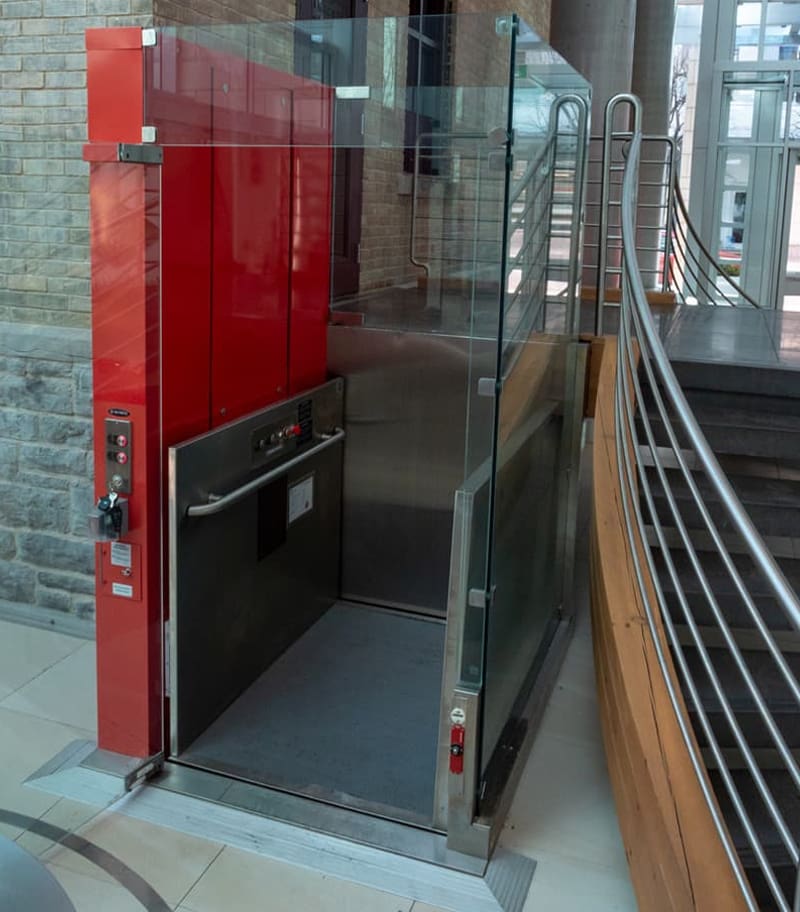 A V-1504 Prestige Vertical Platform Lift by Savaria, featuring red and stainless steel panels with glass sides for wheelchair access, is installed beside a staircase in a modern building. A V-1504 Prestige Vertical Platform Lift by Savaria, featuring red and stainless steel panels with glass sides for wheelchair access, is installed beside a staircase in a modern building.