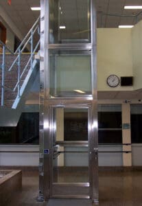 A V-1504 Lux Vertical Platform Lift by Savaria, featuring glass and metal construction, is installed beside a staircase in a building lobby. A clock and sign appear on the pale yellow wall behind the lift.