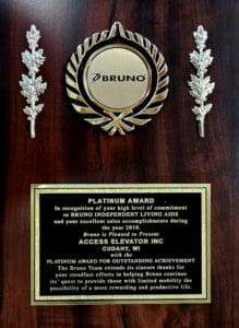 A wooden plaque featuring a gold Bruno medallion with laurel leaves, two silver leaf decorations, and a gold plate inscribed with a Platinum Award for outstanding achievement, awarded by Access Elevator Inc.