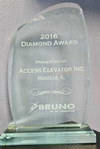 A glass award with engraved text reads: 2016 Diamond Award presented to Access Elevator Inc, Roselle, IL. BRUNO for your Independence. The award has a curved shape and sits on a glass base.
