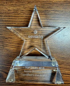 A clear, star-shaped glass award on a wooden surface. The award is engraved with Savaria, TOP SALES 2018, and Access Elevator Inc., certified 5-star dealer.