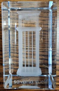 A clear rectangular glass block with an etched 3D image of a columned structure inside, placed on a wooden surface. The text Savarta vuelif is visible at the bottom of the block.