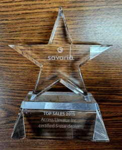 A clear glass star-shaped award sits on a wooden surface. Engraved text reads: savaria. TOP SALES 2019. Access Elevator Inc. certified 5-star dealer.