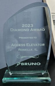 A glass award with the inscription: 2023 Diamond Award Presented to Access Elevator Roselle, IL and the Bruno company logo, displayed on a dark surface against a gray wall.