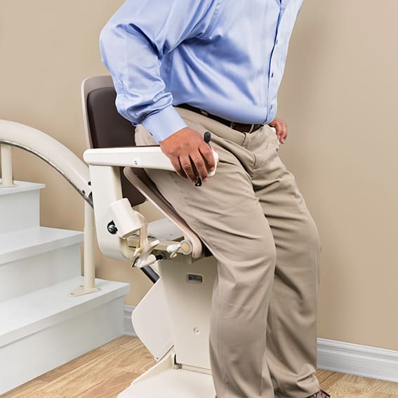 A person in khaki pants and a blue shirt sits at the bottom of a staircase on a Freecurve Curved Stair Lift by Handicare, preparing to use this residential lift for safe, easy access to the upper floor.