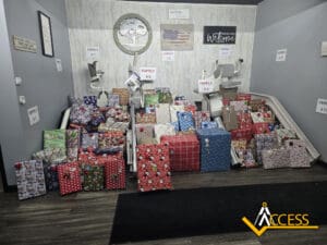 A variety of wrapped gifts are stacked on and around two stair lifts under signs labeled Family #5 and Family #6 in a gray room. A yellow “ACCESS” logo is visible in the bottom right corner.