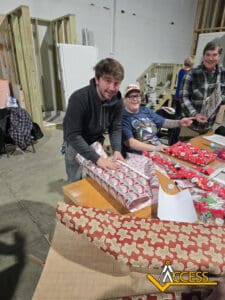 Two smiling people wrap Christmas gifts at a table covered with wrapping paper and supplies in a warehouse-like space. Another person stands nearby. The ACCESS logo is visible in the bottom right corner.