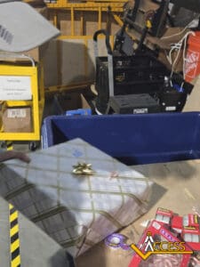 A person holds a wrapped gift with a gold bow over a table with candy and an Access logo, in a warehouse setting with shelves, carts, and equipment in the background.