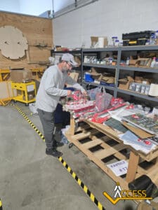 A man wearing a gray hat and shirt wraps holiday gifts at a wooden table in a warehouse. Gifts, wrapping paper, and supplies are spread out; shelving with boxes and bins lines the wall. The ACCESS logo appears in the corner.