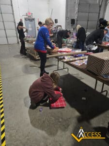 Several people are wrapping gifts on tables in a large warehouse. A child in the foreground kneels to wrap a red box. The ACCESS logo is visible in the bottom right corner.