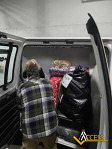 A person wearing a plaid shirt loads large bags and wrapped gifts into the back of a white van. The ACCESS logo is visible in the bottom right corner.