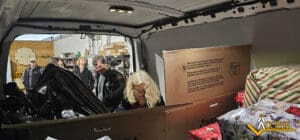 Several people stand at the back of a van filled with large boxes and wrapped gifts, preparing or organizing donations inside a warehouse. The ACCESS logo is visible in the corner.