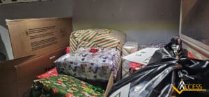 A collection of wrapped gifts and boxes, including striped, snowman, and green-themed wrapping paper, piled together in a small space. A black plastic bag and ACCESS logo are also visible in the foreground.