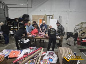 A group of people wrap gifts on tables in a warehouse with shelves, supplies, and wrapping paper rolls. Some wear ACCESS shirts; the ACCESS logo is visible in the lower right corner.