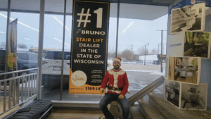 A man in a Santa hat and festive sweater sits on a stair lift inside a showroom. A large sign behind him reads #1 Bruno Stair Lift Dealer in the State of Wisconsin with snow visible outside the windows.