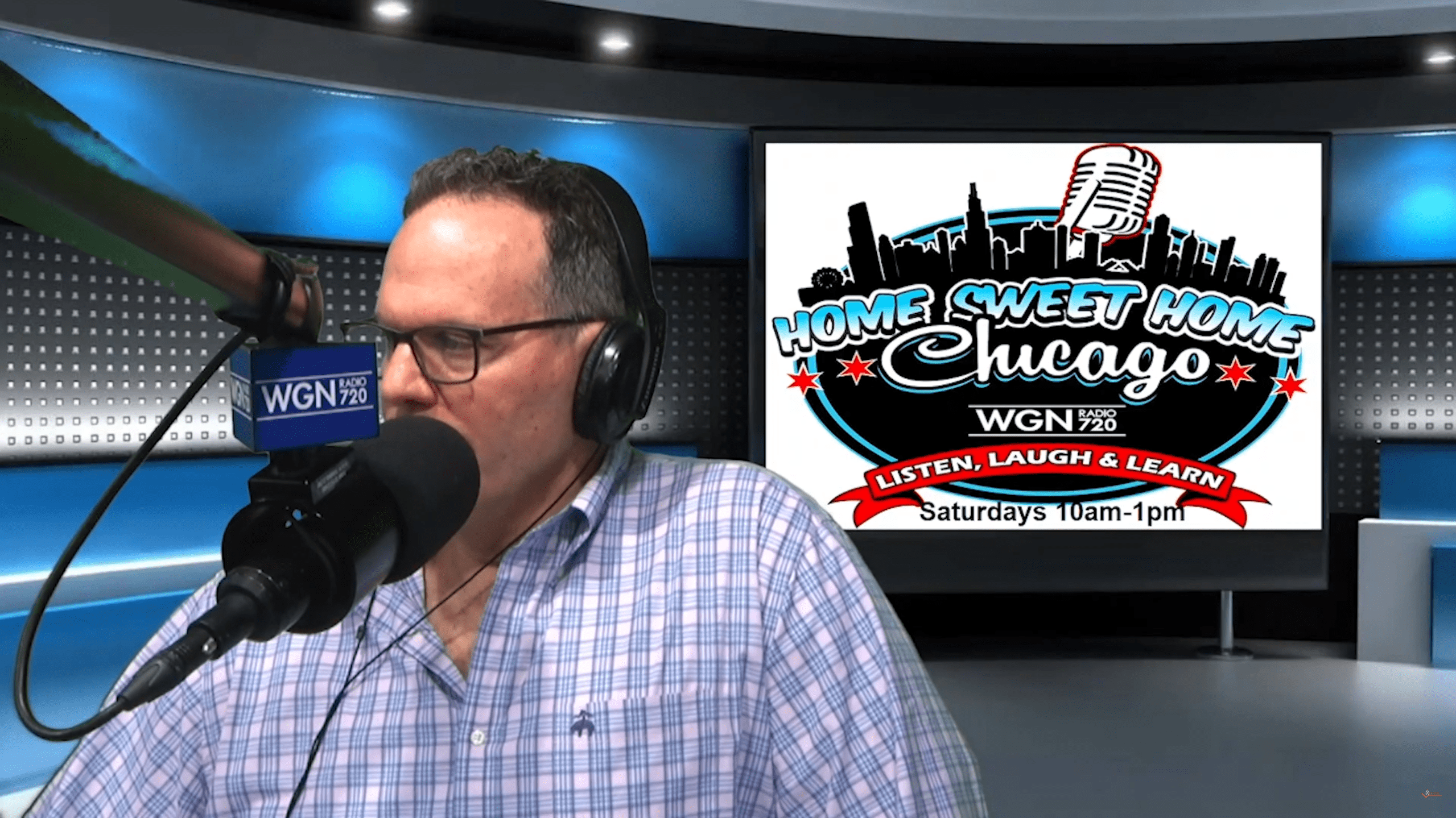 A man wearing glasses and headphones speaks into a WGN 720 microphone in a radio studio. Behind him, a screen displays the Home Sweet Home Chicago show logo and its schedule: Saturdays 10am–1pm.