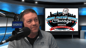 A man wearing a gray jacket speaks into a microphone in a radio studio. Behind him is a screen displaying the Home Sweet Home Chicago show logo and broadcast details for WGN Radio 720, airing Saturdays from 10am to 1pm.