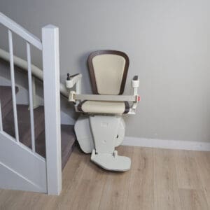 A Freecurve Curved Stair Lift by Handicare with a beige seat and brown trim is installed at the base of a carpeted staircase beside a white railing on light wood flooring.