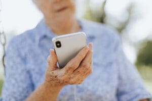 An older adult wearing a blue patterned shirt holds a smartphone in one hand, looking at the screen. The background is blurred, suggesting an outdoor setting.