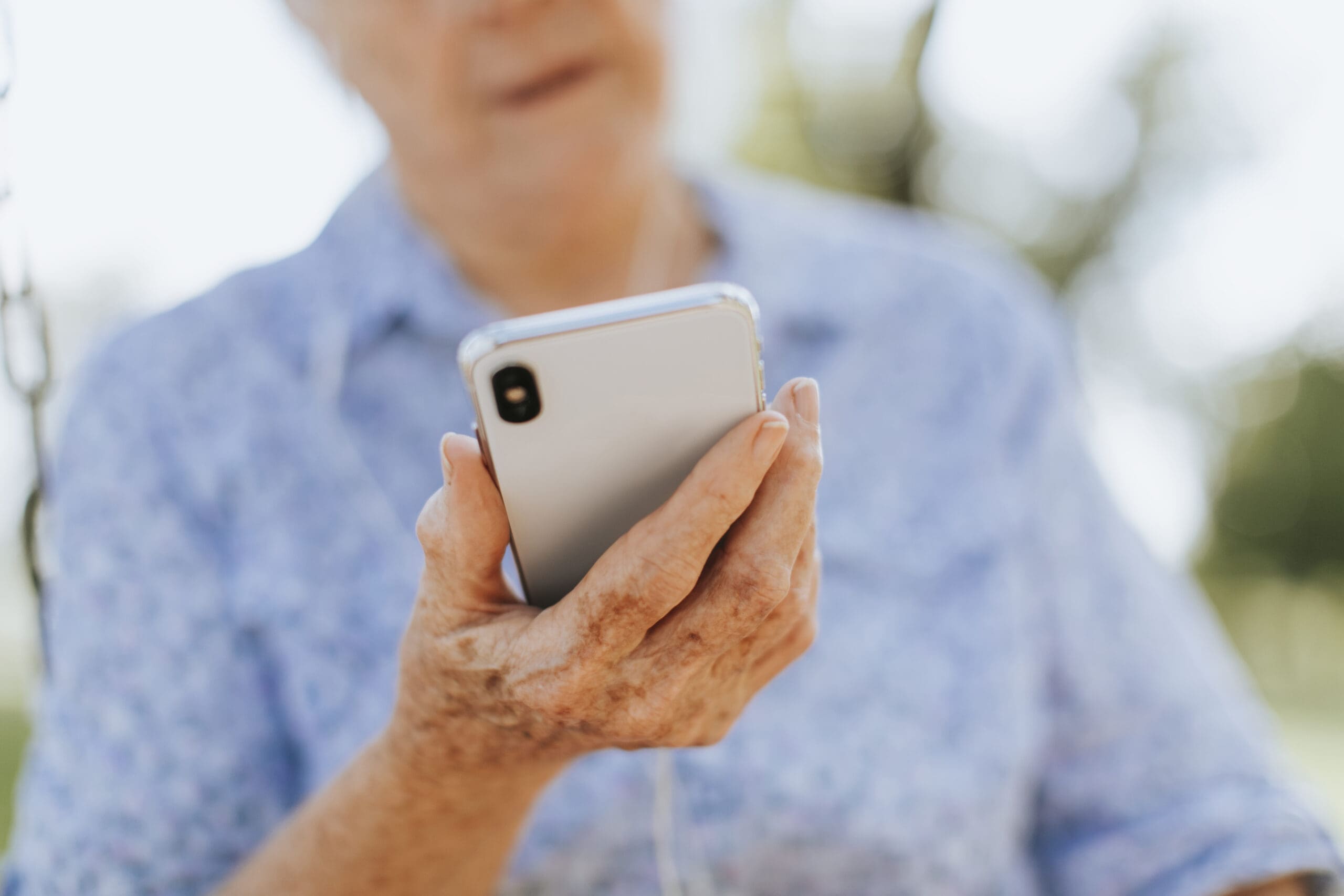 An older adult wearing a blue patterned shirt holds a smartphone in one hand, looking at the screen. The background is blurred, suggesting an outdoor setting.